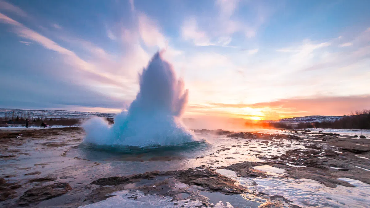 Geyser erupting with water shooting upward during a colorful sunset over rocky terrain.