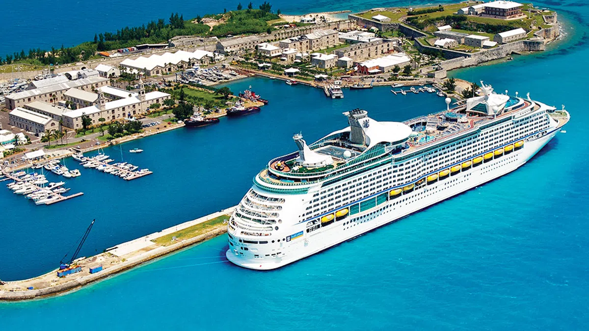 Large white cruise ship docked near a tropical island with clear blue waters and marina boats.