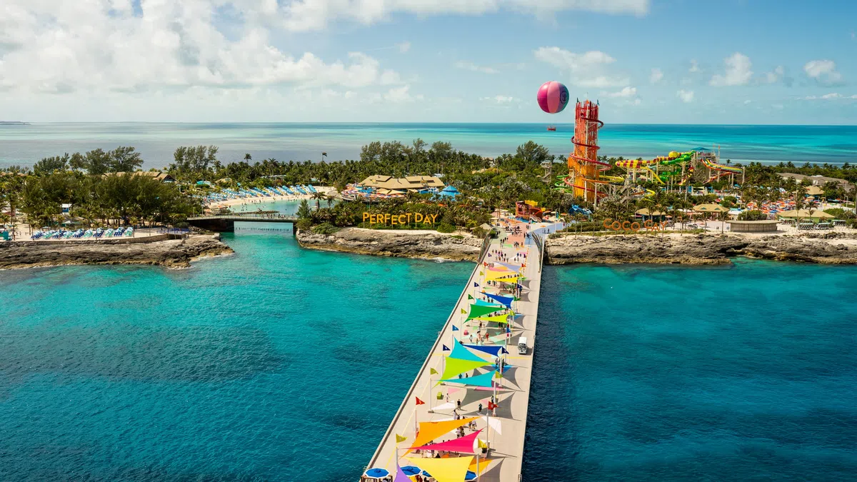 Aerial view of a colorful pier leading to a tropical island resort with pools, water slides, and clear turquoise ocean.