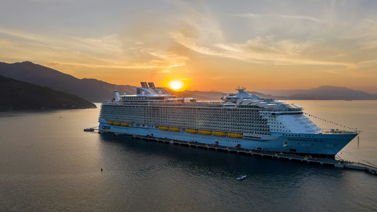 Large cruise ship docked at sunset with mountains and calm sea in the background under a colorful sky