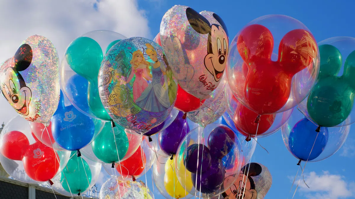 Colorful Disney-themed balloons including Mickey Mouse and princesses floating against a blue sky with clouds.