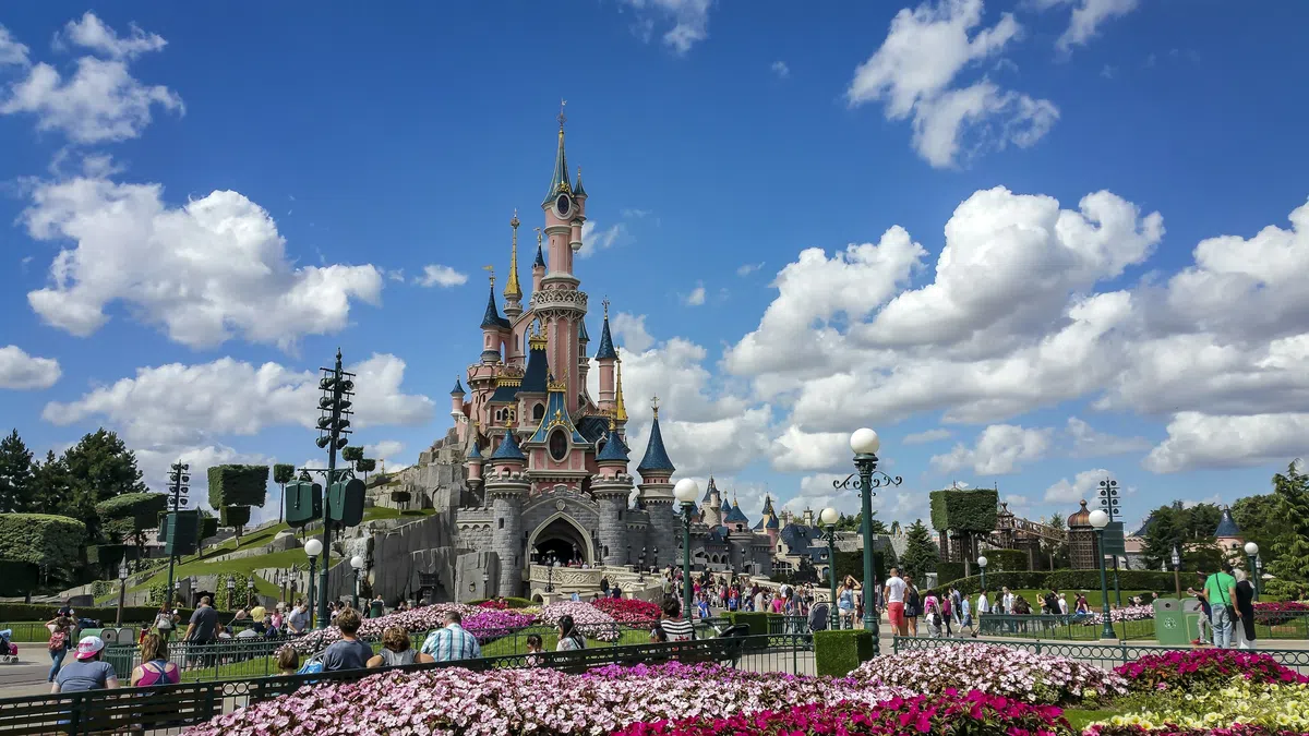Disneyland Paris castle with pink towers and blue spires surrounded by colorful flowers and visitors under a bright blue sky.