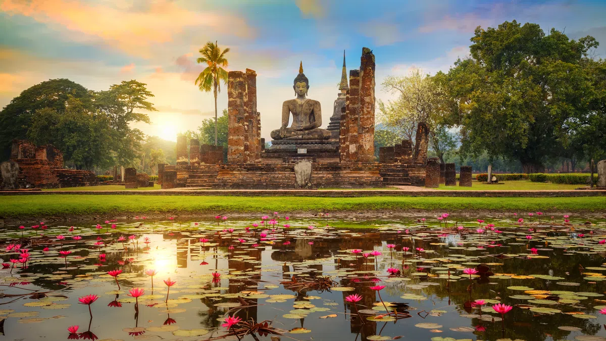 Sunset over ancient Buddha statue ruins with pink lotus flowers blooming in a reflective pond in Sukhothai.