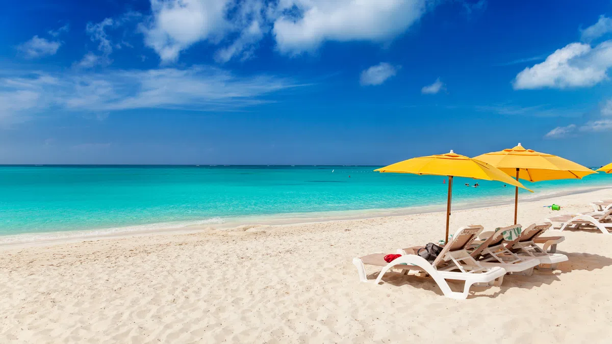 White sandy beach with turquoise water, yellow umbrellas, and empty lounge chairs under a partly cloudy blue sky.