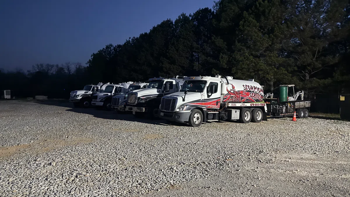 Fleet of white service trucks parked on gravel lot with trees in the background at dusk.