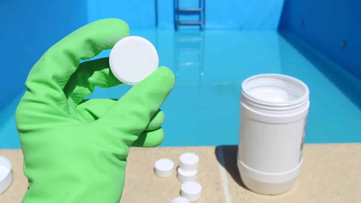 Hand in green glove holds a white chlorine tablet near a clean swimming pool for water treatment.