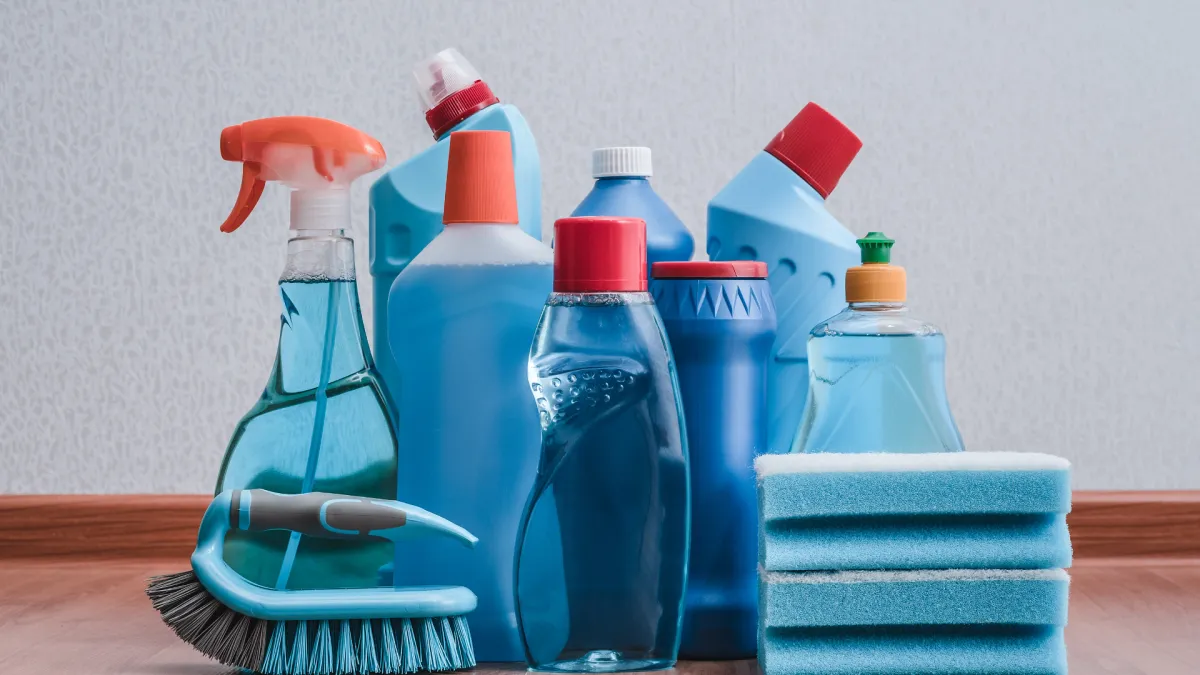 Assortment of blue and red cleaning bottles, brushes, and sponges arranged on wooden floor against white wall.