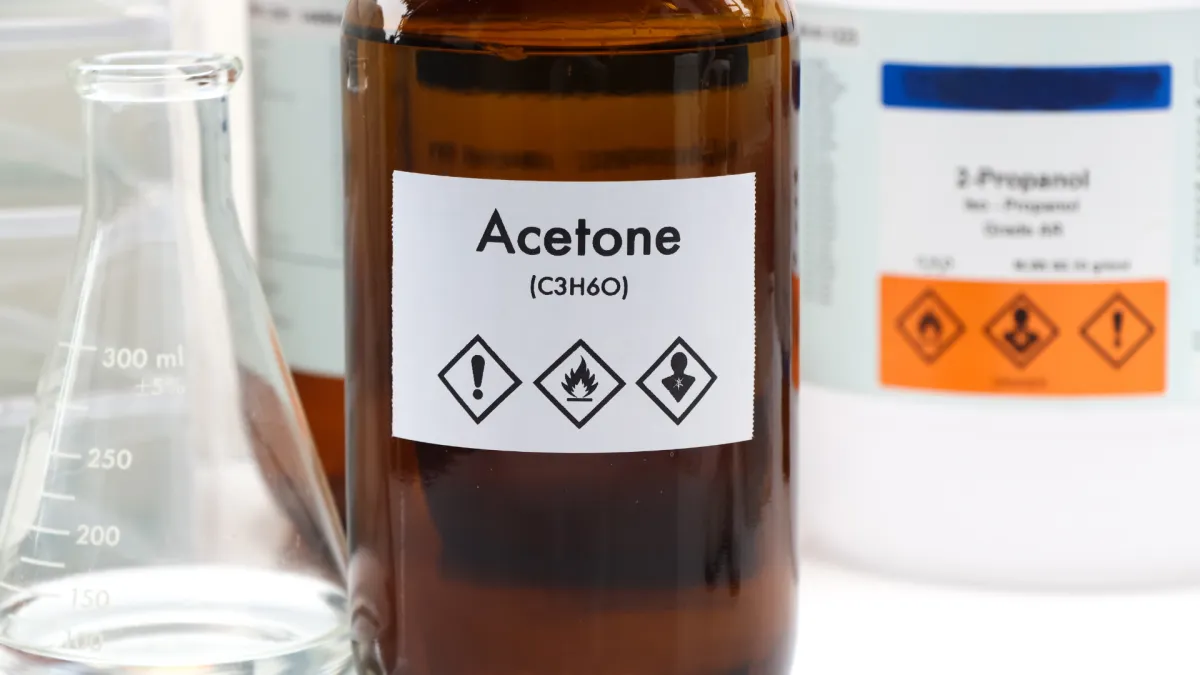 Brown bottle labeled acetone with hazard symbols alongside laboratory glassware and chemical containers.