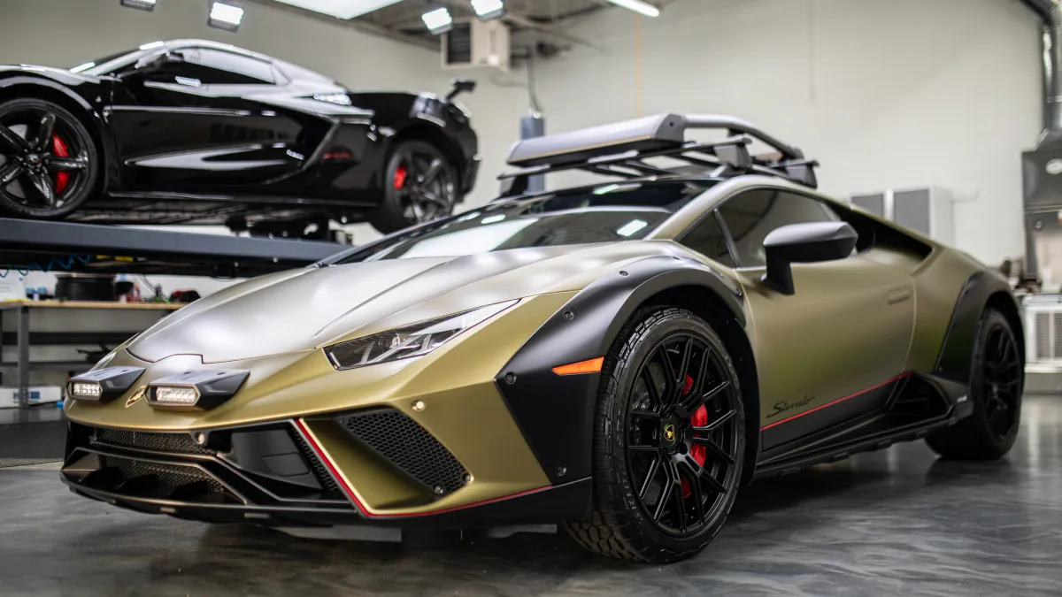 Matte gold Lamborghini Huracan with black accents and roof rack in a modern garage setting.