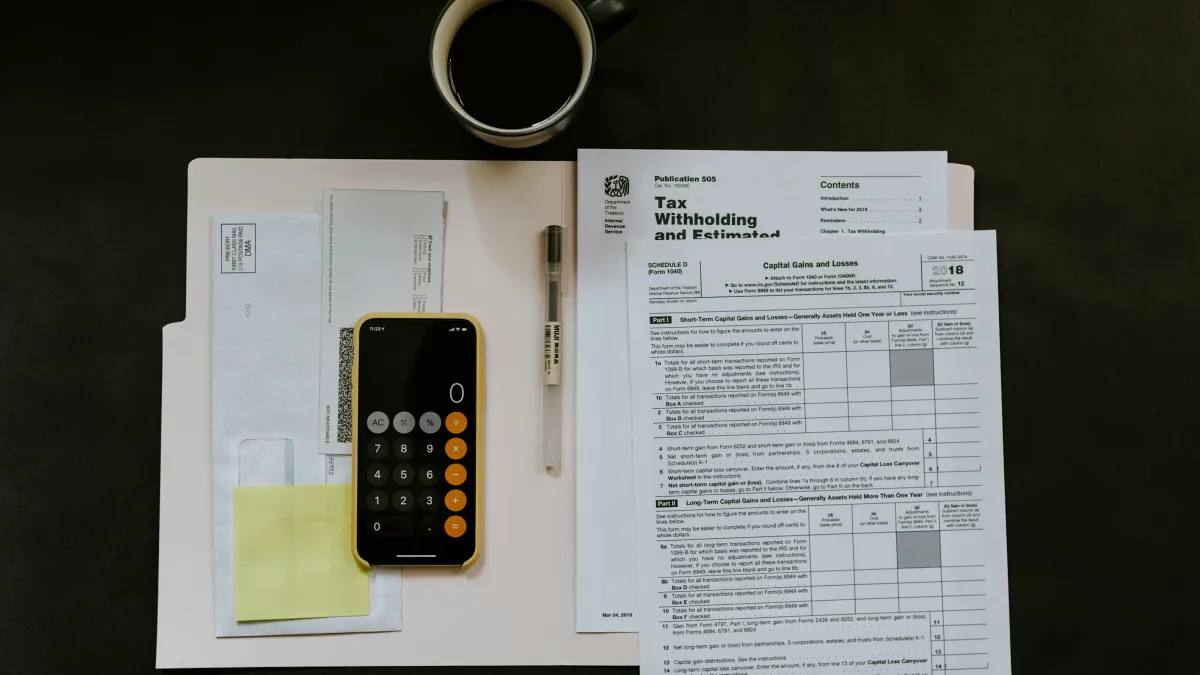 Overhead view of tax forms, a calculator app on a smartphone, envelopes, pen, and a coffee cup on a dark surface