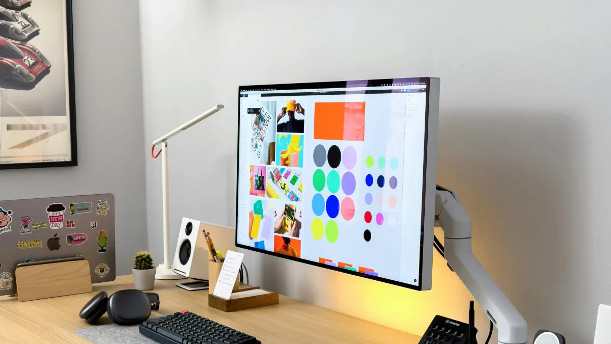 Modern clean desk setup with monitor displaying colorful designs, keyboard, mouse, laptop with stickers, and desk lamp.