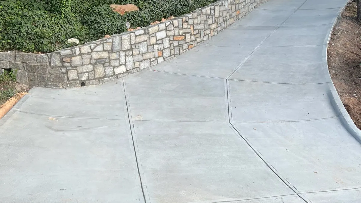 Concrete driveway sloping upward alongside stone retaining wall and green wooded hillside near wooden house.
