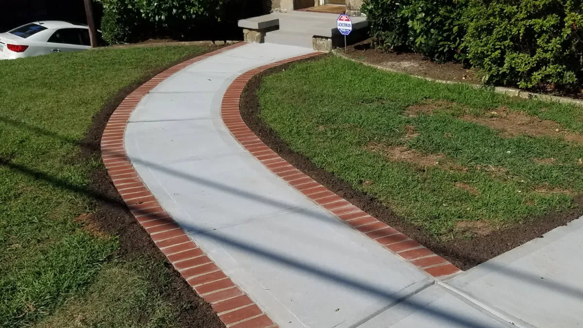 Curved white concrete walkway with red brick border leading to a house entrance with stone and brick facade.