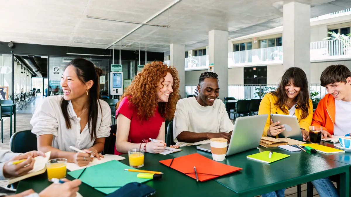 Group of diverse young adults studying and collaborating around a table with laptops, notebooks, and drinks in a modern indoor space