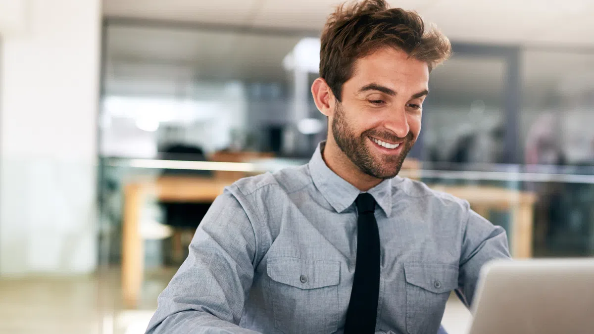 Smiling businessman in gray shirt and black tie working on laptop in bright modern office environment