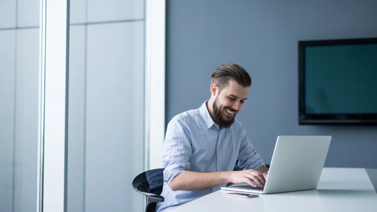 Smiling man in striped shirt working on a laptop at a white desk in a modern office with large windows.