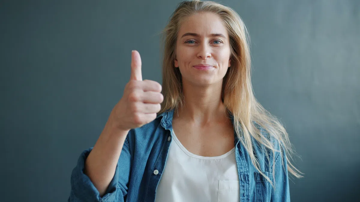 Smiling young woman wearing a denim jacket giving a thumbs up against a plain gray background