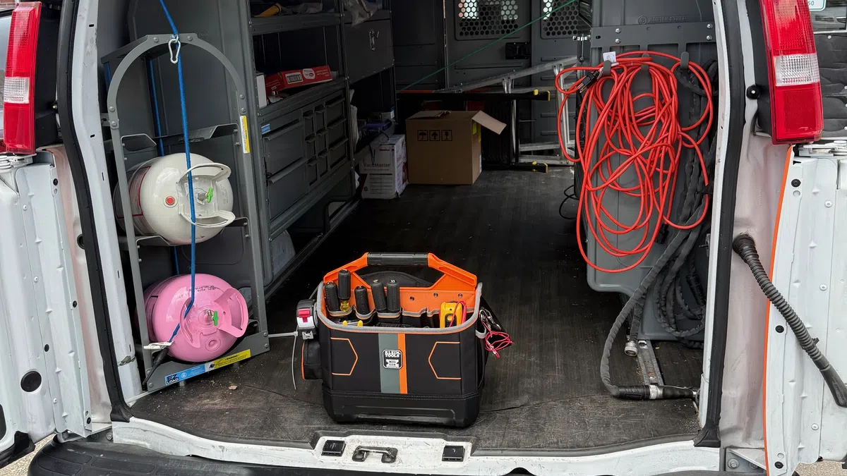 Open back of a work van showing toolboxes, safety helmets, extension cords, and ladders on top.