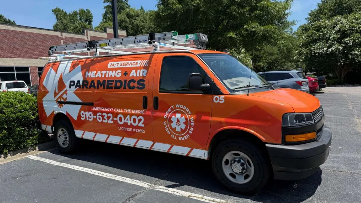 Orange Heating + Air Paramedics service van parked in a lot with contact number and ladder on roof.