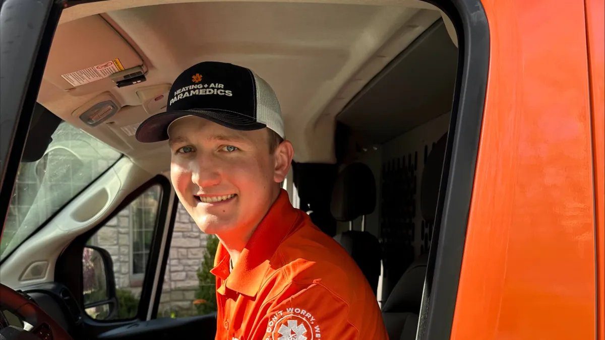 Smiling paramedic in orange uniform sitting in driver's seat of an orange van with branded cap.