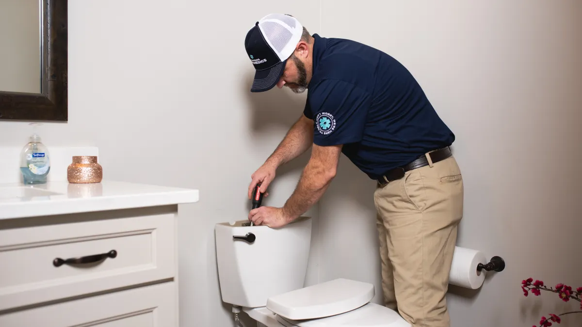 Plumber fixing toilet tank with screwdriver in a clean bathroom wearing uniform and cap.