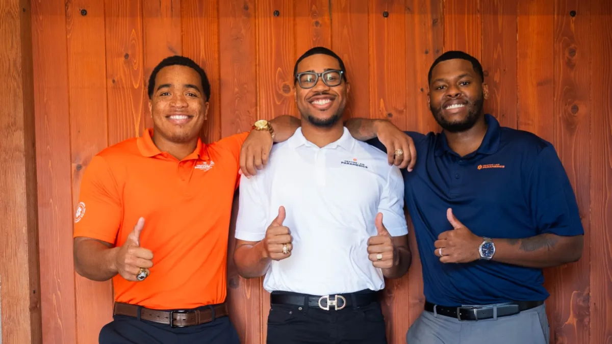 Three men smiling and giving thumbs up while standing and leaning against a wooden wall indoors.
