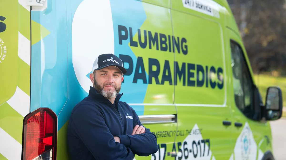 Plumbing paramedics technician standing with arms crossed next to a branded service van outdoors.
