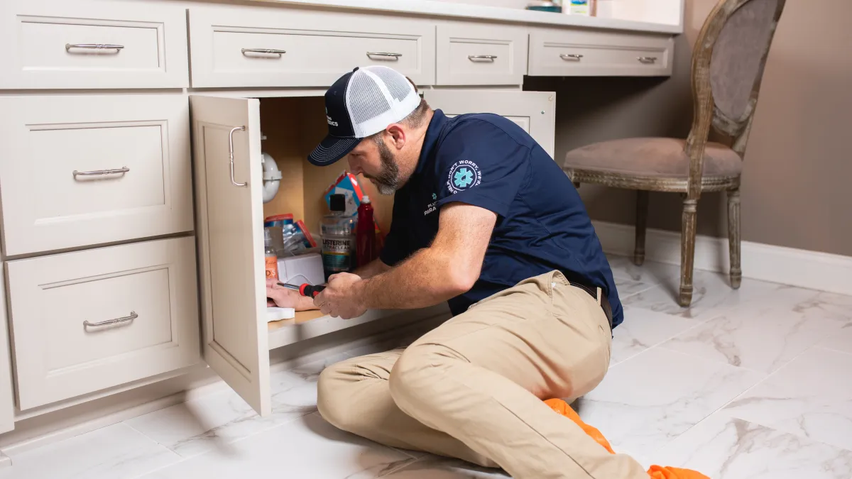 Plumber in uniform kneeling on marble floor fixing pipes under bathroom sink cabinet.