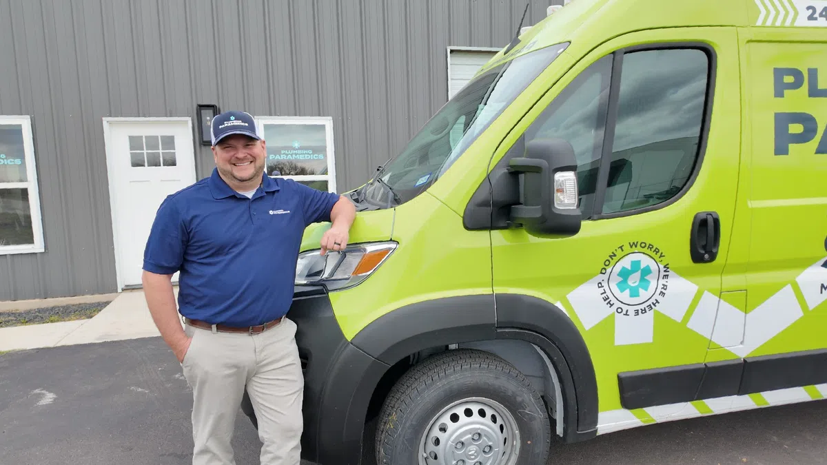 Smiling man in blue shirt and hat leans on bright green emergency paramedic van outside gray building.