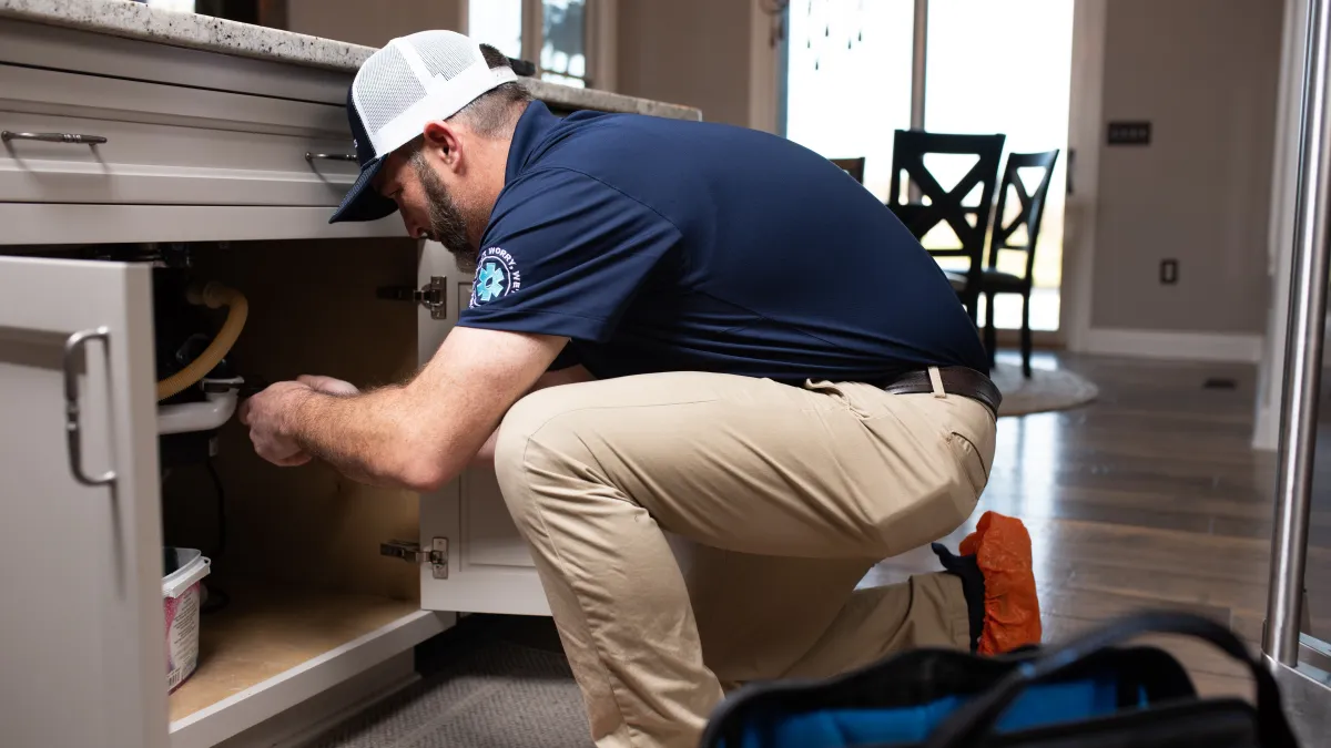 Technician in cap and uniform repairing plumbing under kitchen sink in modern home interior.