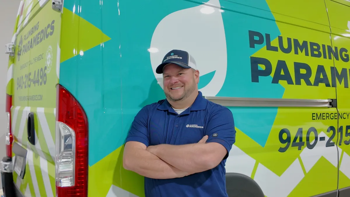 Smiling plumber in uniform standing with arms crossed beside Plumbing Paramedics van with contact info visible
