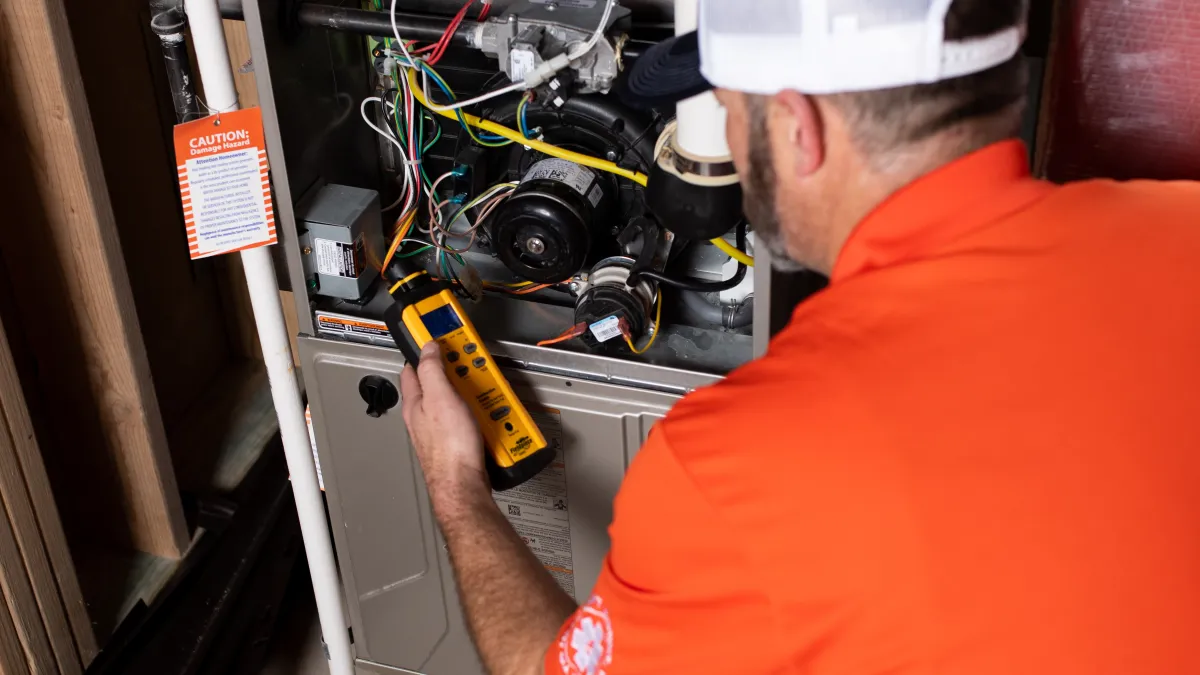 Technician in orange shirt inspecting and testing an open furnace unit with a handheld diagnostic tool indoors.