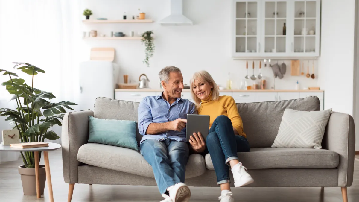 Smiling senior couple sitting on sofa using tablet in bright modern living room with cozy decor