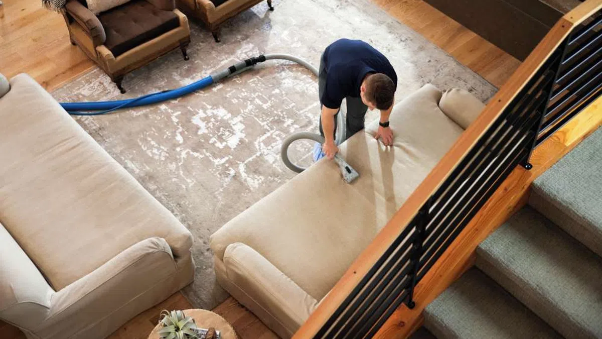 Man cleaning beige sofa upholstery in living room with vacuum tool during professional furniture cleaning service.
