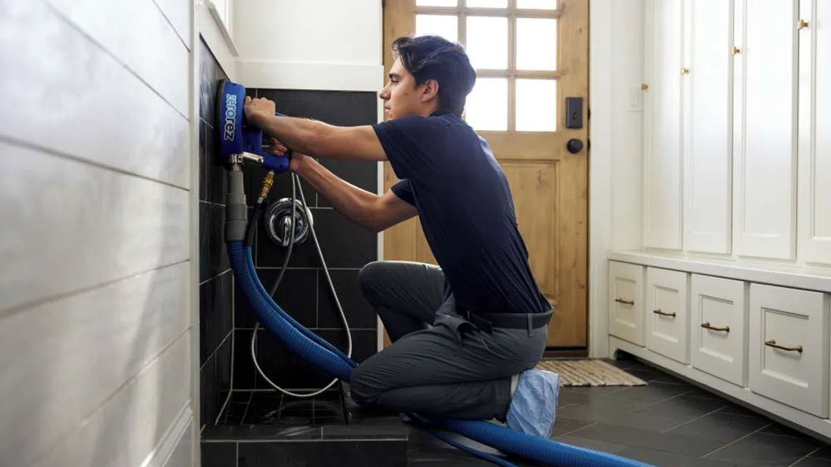 Technician kneeling on black tiled laundry room floor operating a blue carpet cleaning machine hose
