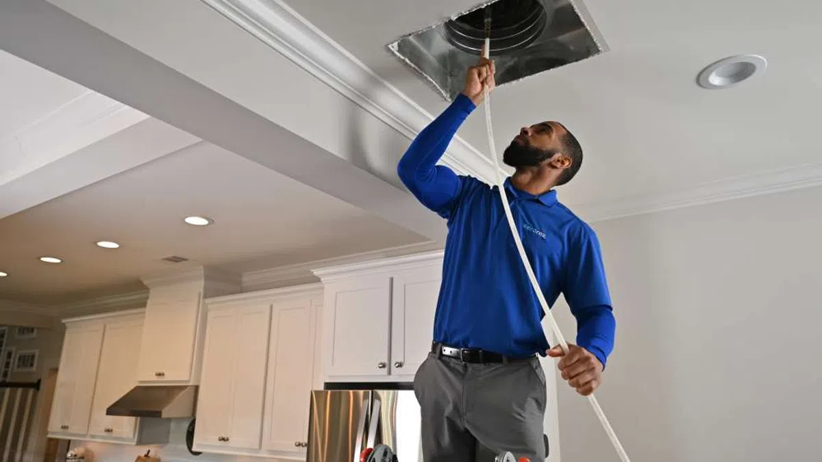 Technician in blue shirt inspecting air duct on ceiling inside a modern kitchen, standing on a ladder