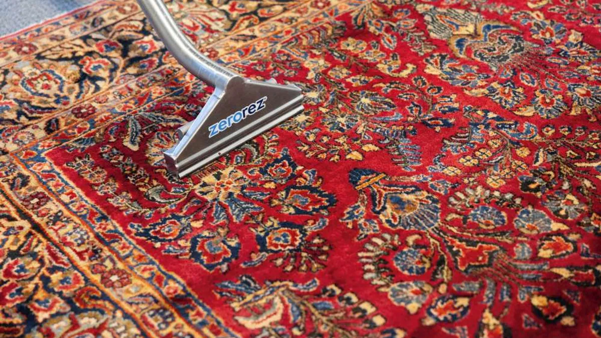 Close-up of a red patterned rug being cleaned with a Zerorez branded carpet cleaning tool with overlaid text reading Zerorez