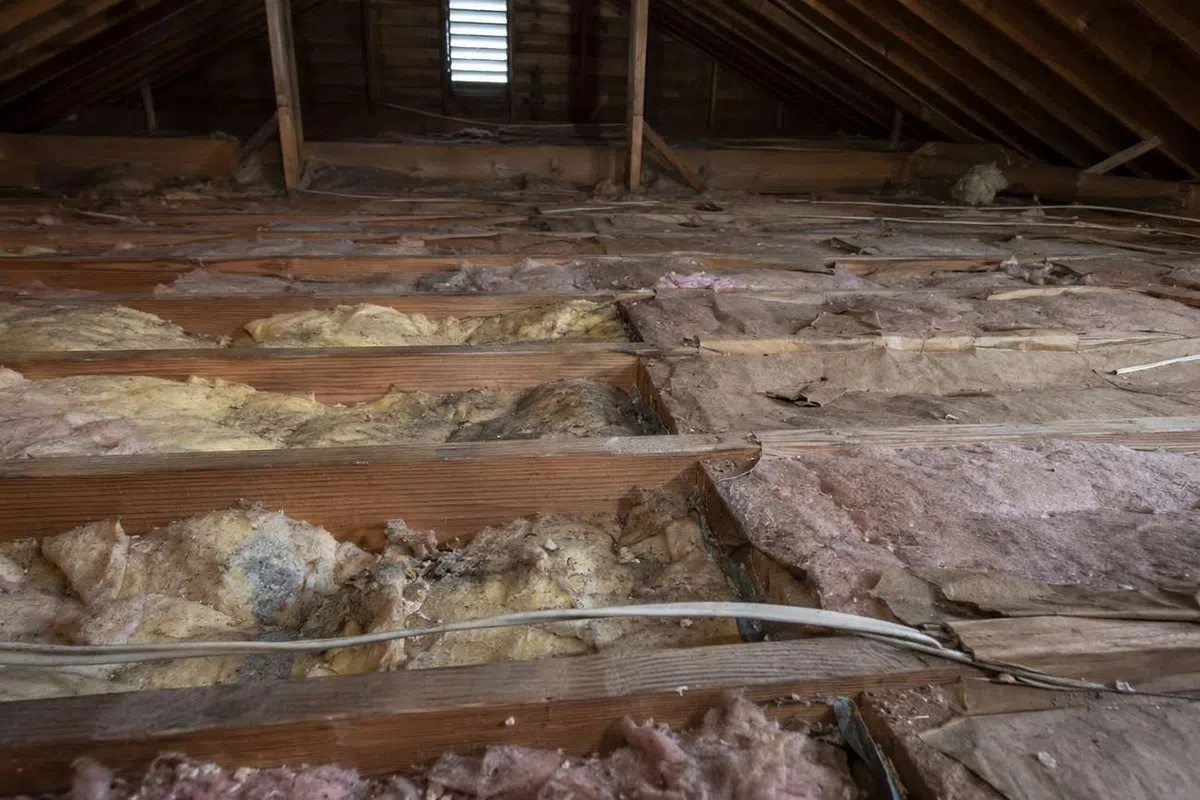 Attic space with exposed wooden beams and old fiberglass insulation installed unevenly between joists.