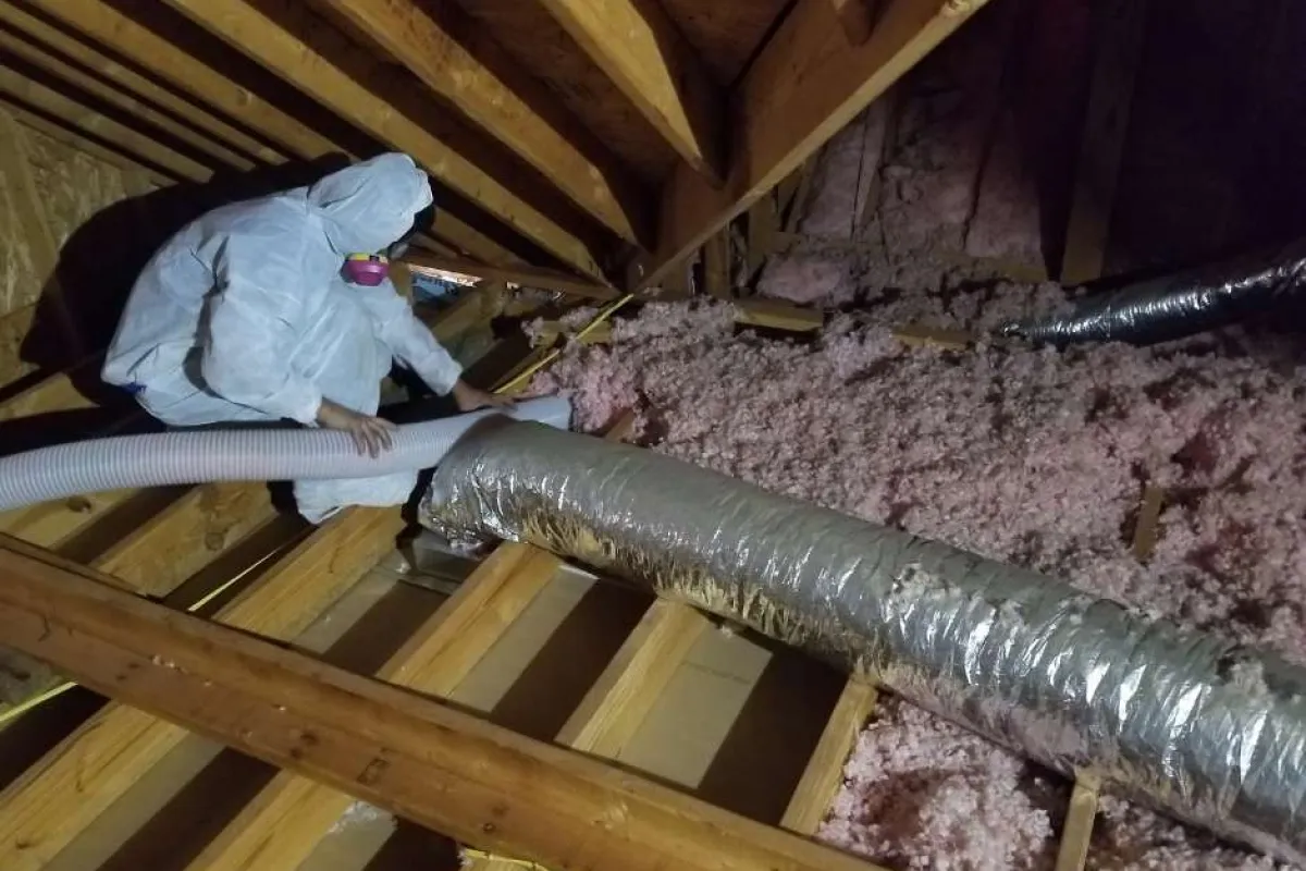 Worker in protective suit installing insulation in attic with pink fiberglass and ductwork under wooden beams.