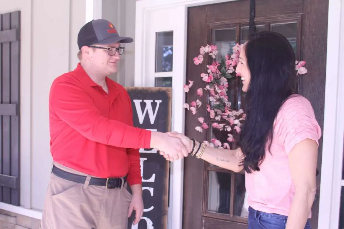 Man in red shirt shaking hands with woman in pink shirt at home entrance with welcome sign and flowers.