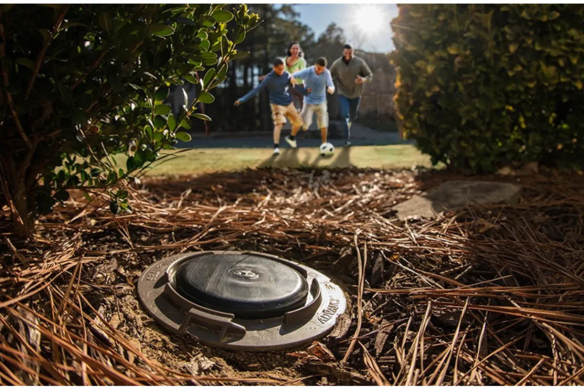 Outdoor irrigation valve surrounded by mulch and bushes with a family playing soccer in the background.