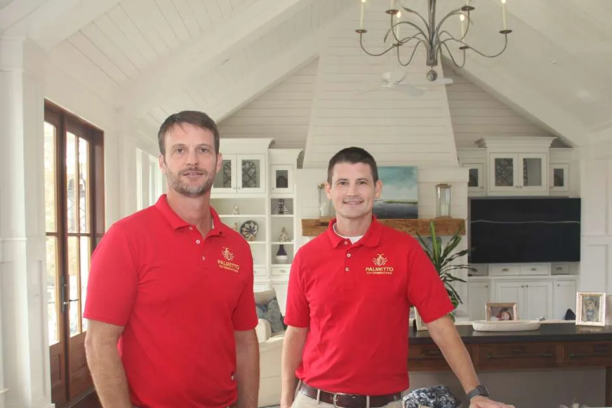Two men wearing red Palmetto pest control polo shirts standing in a modern kitchen with white cabinetry and a marble island.