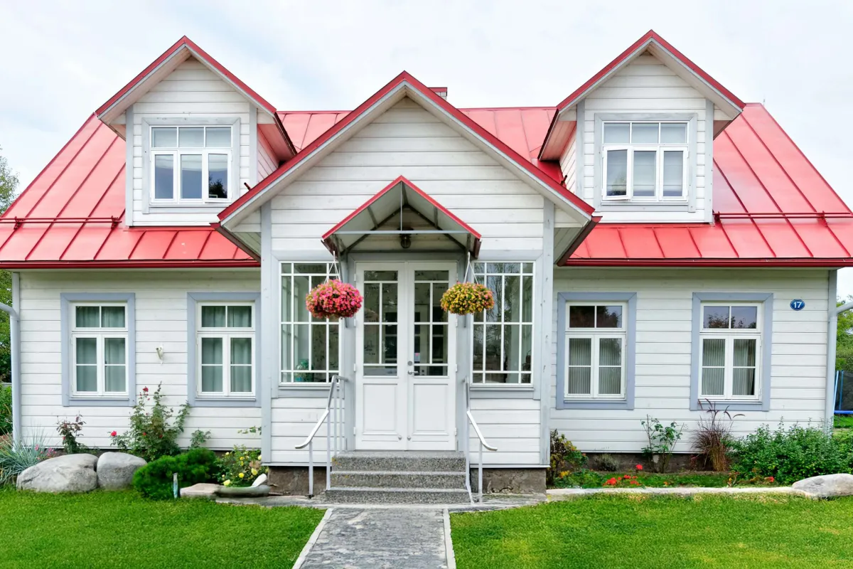 White house with red metal roof, front porch, hanging flower pots, and green lawn with stone pathway.