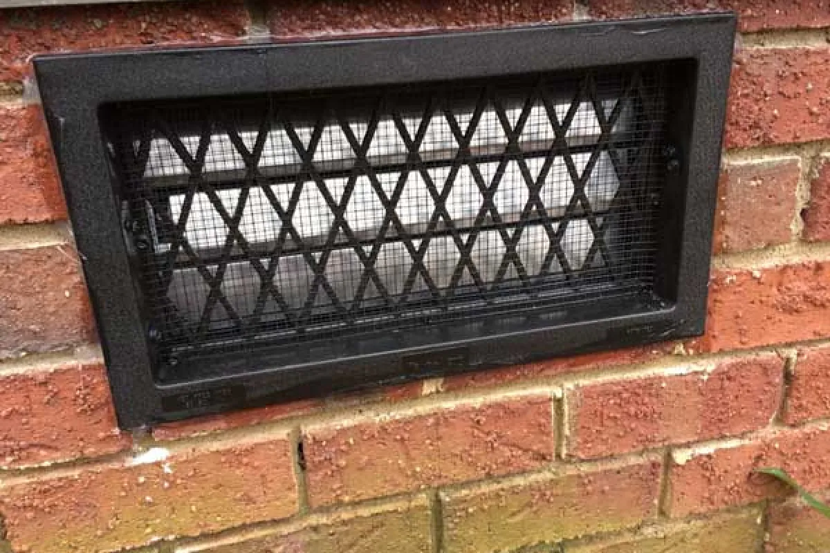 Black metal vent cover with cross-hatch design installed on red brick wall exterior near ground level.