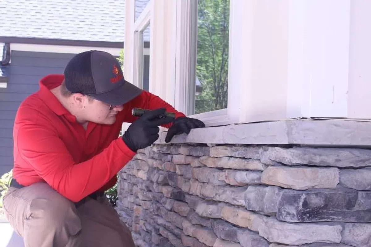 Man in red shirt inspecting stone wall exterior of house with flashlight during daytime.