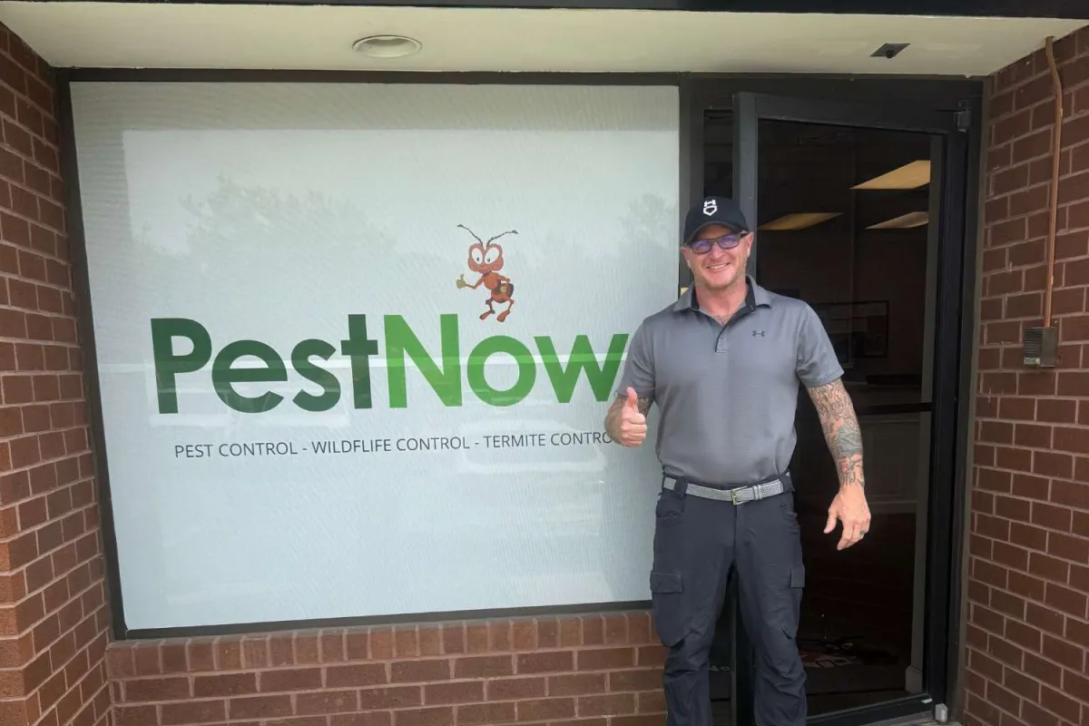 Man in gray uniform giving thumbs up outside PestNow pest control office with logo on window.
