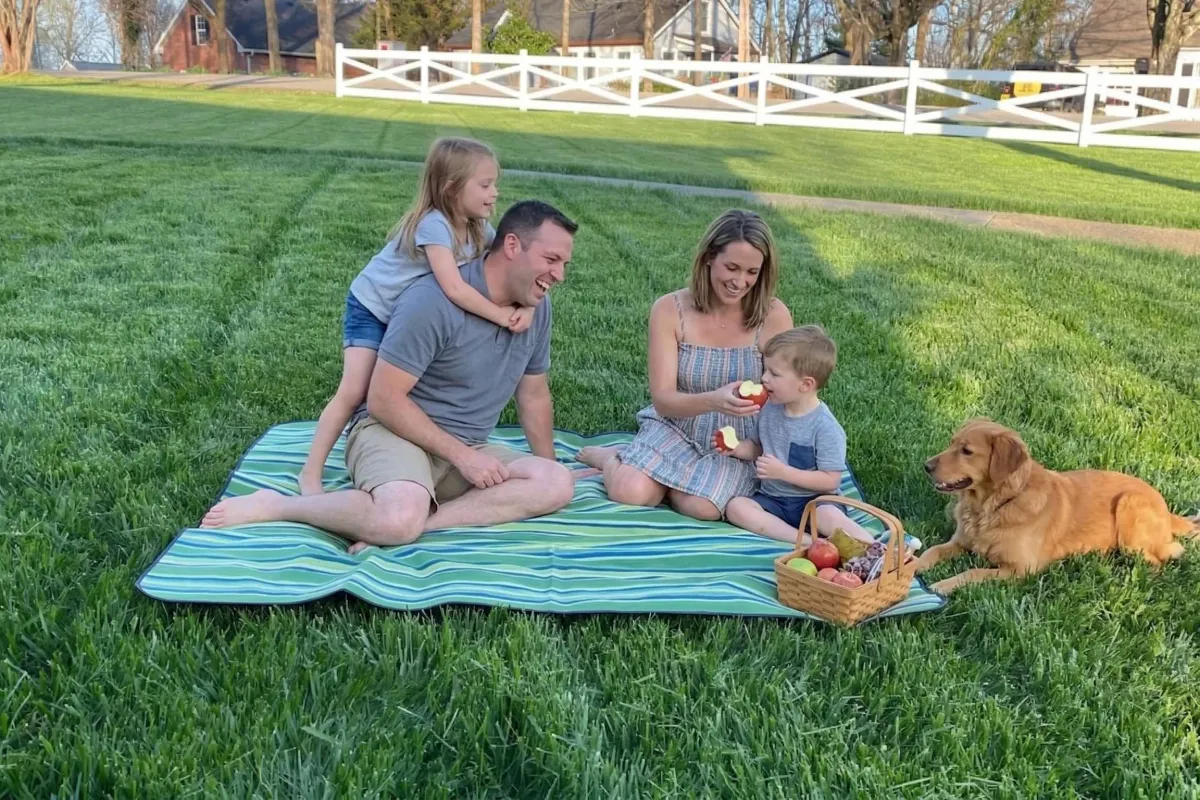 Family of four and golden retriever enjoying picnic on green grass with basket of apples on a sunny day