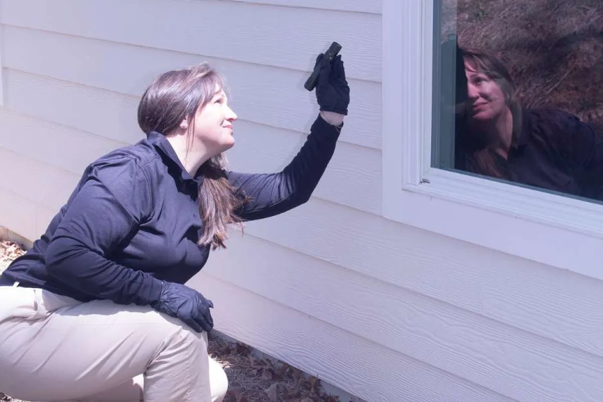 Woman in black shirt and gloves inspecting house exterior near window during daylight.
