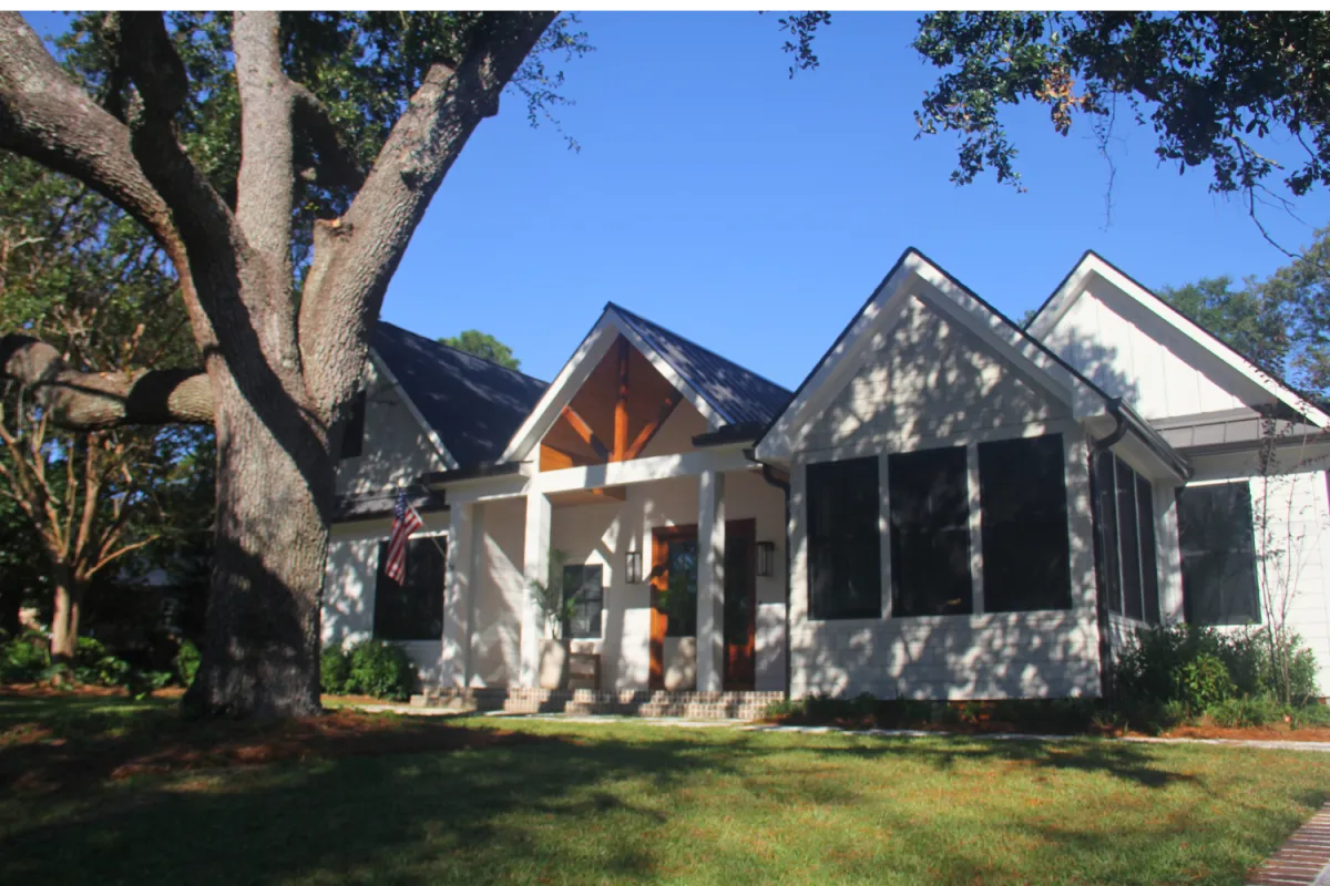 Modern white house with gable roof, large front porch, tall tree, and green lawn under clear blue sky.