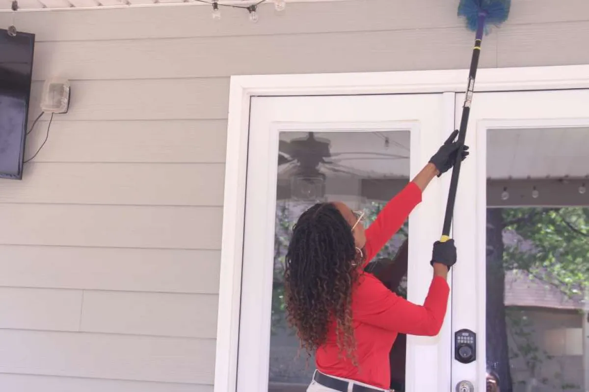 Woman cleaning ceiling fan blades on porch with a blue duster, wearing red shirt and gloves near glass door.